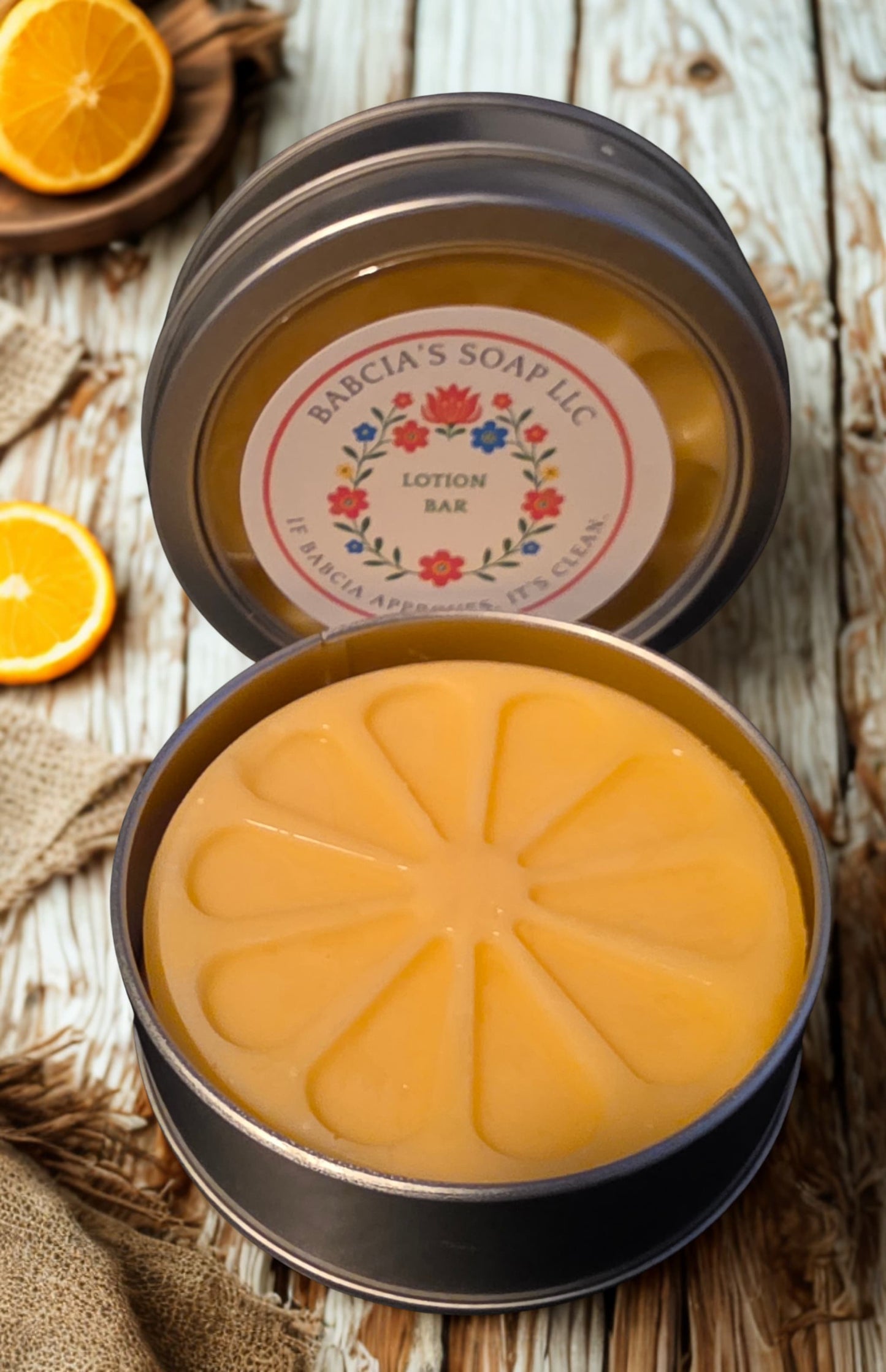 An open round metal tin of Bacia's Soap LLC Lotion Bar, showing the orange-colored bar with a segmented pattern, set on a rustic wooden surface with sliced oranges.