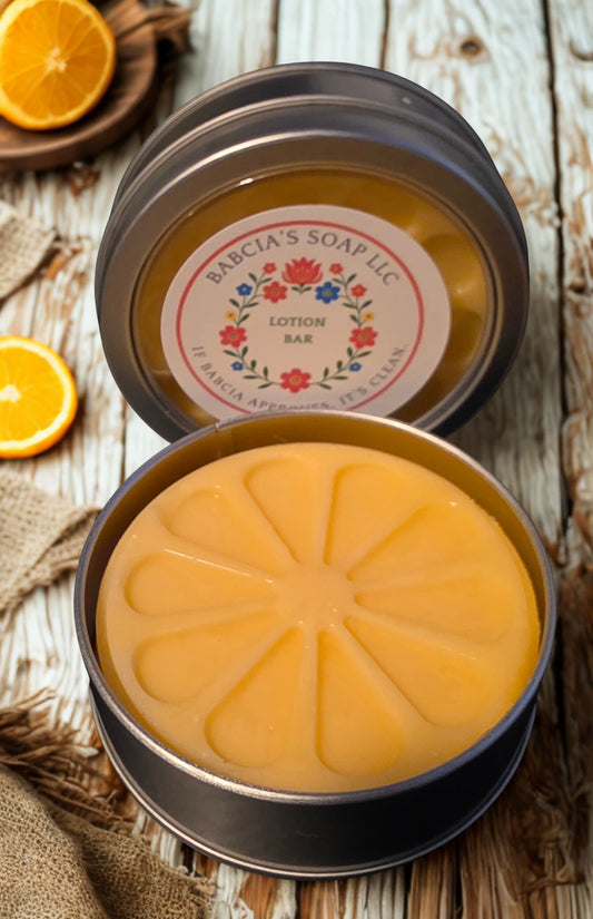 An open round metal tin of Bacia's Soap LLC Lotion Bar, showing the orange-colored bar with a segmented pattern, set on a rustic wooden surface with sliced oranges.