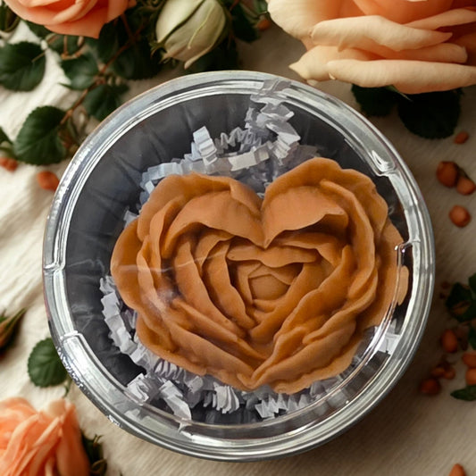 A heart-shaped brown wax melt or soap piece displayed inside a clear round plastic container with shredded white paper filler, surrounded by peach roses and small orange beads on a beige fabric surface.